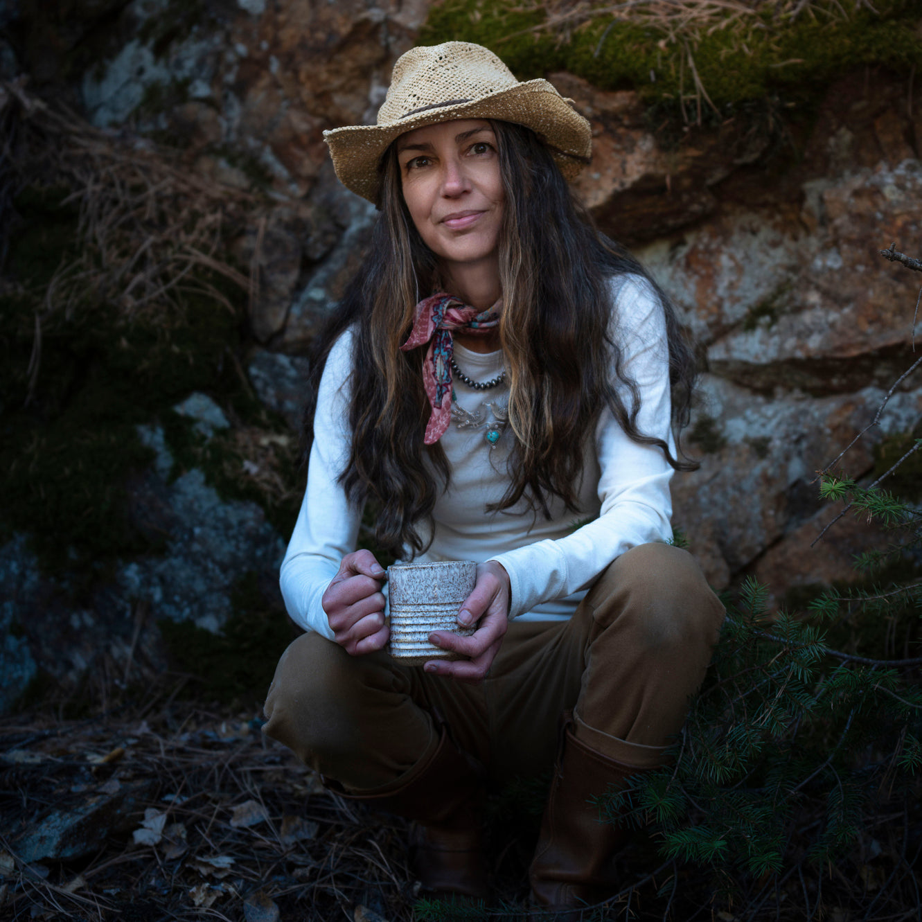 Amy in a straw hat and brown pants holding a ceramic mug in a natural setting.