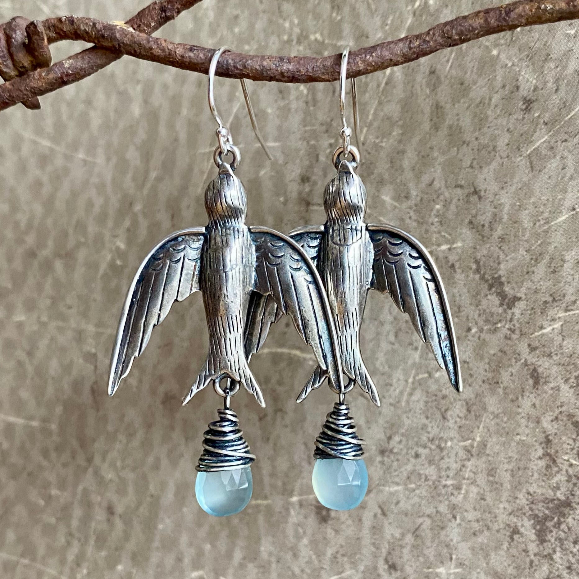 Silver bird-shaped earrings with blue teardrop charms hanging from a branch.