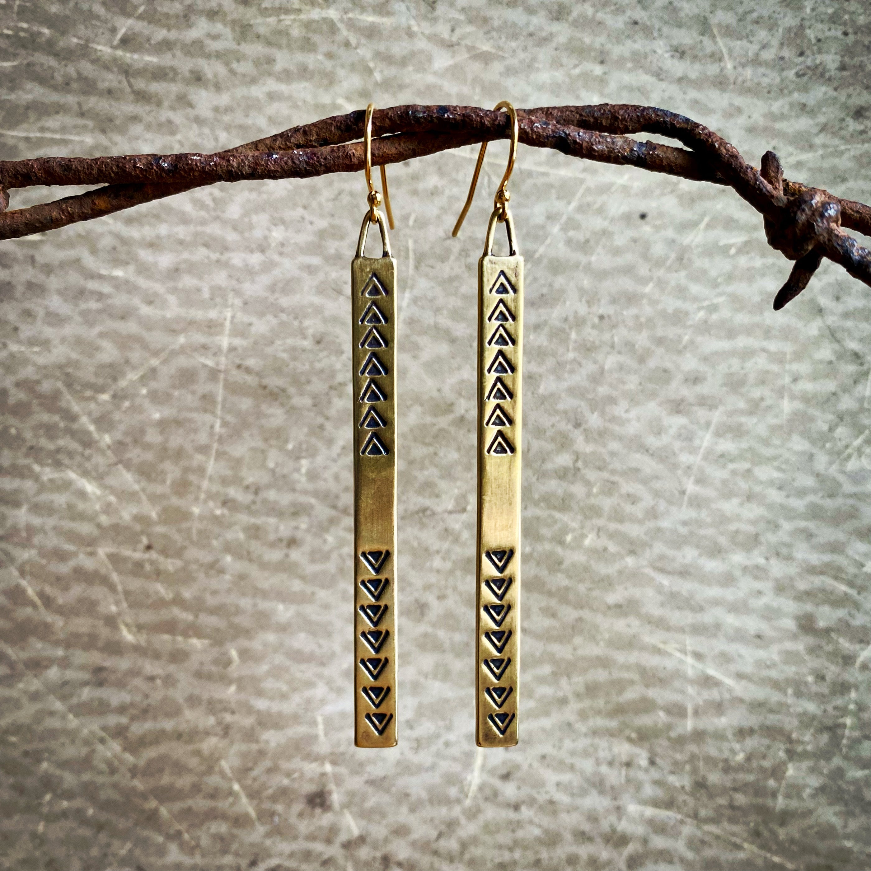 Pair of gold earrings with triangle patterns hanging on a branch.