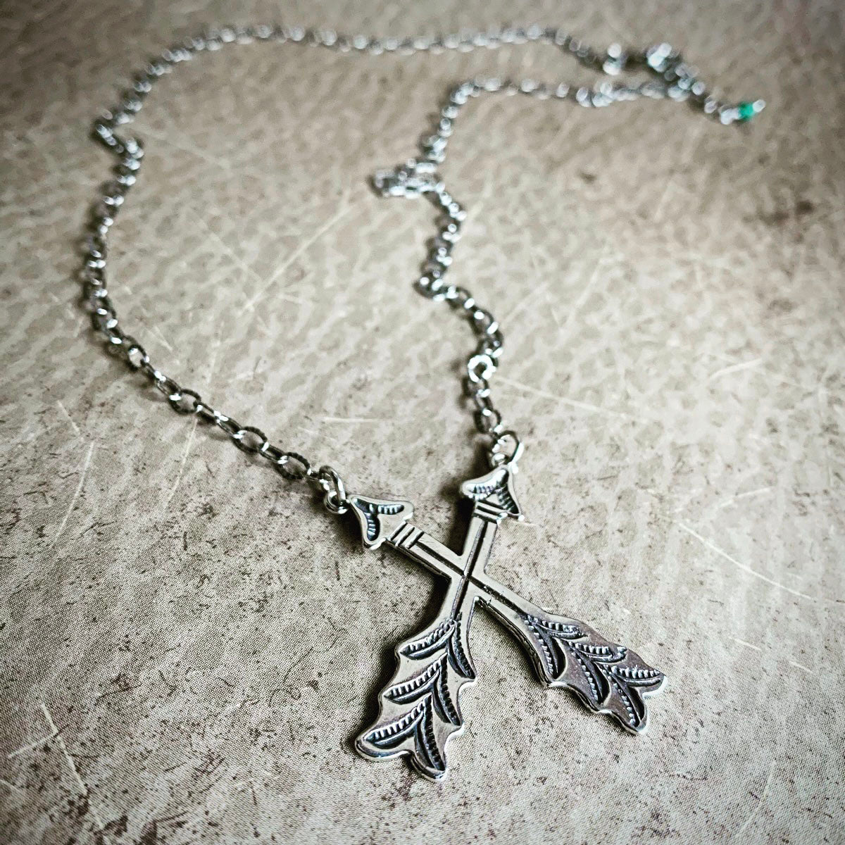 Silver cross arrows-shaped pendant on a chain against a textured beige background