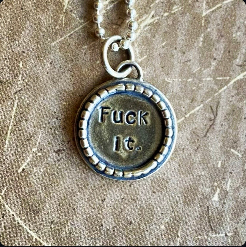 Silver necklace with a pendant that says 'F**k it.' on a textured surface