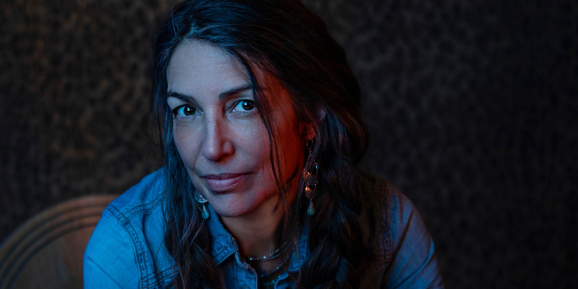 Amy with dark hair and a denim jacket sitting against a textured wall.